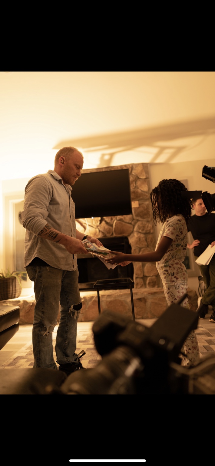Two performers exchange papers while cameras and crew work around them.