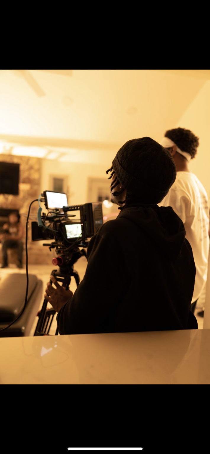 Camera operator films from behind the camera in a warmly lit room.