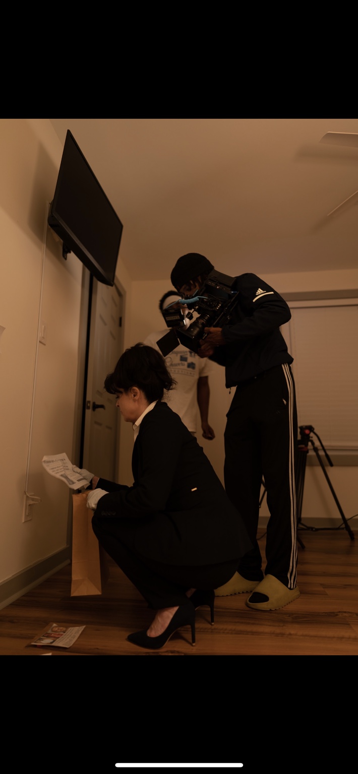 A camera operator films a performer crouching beside a wall.