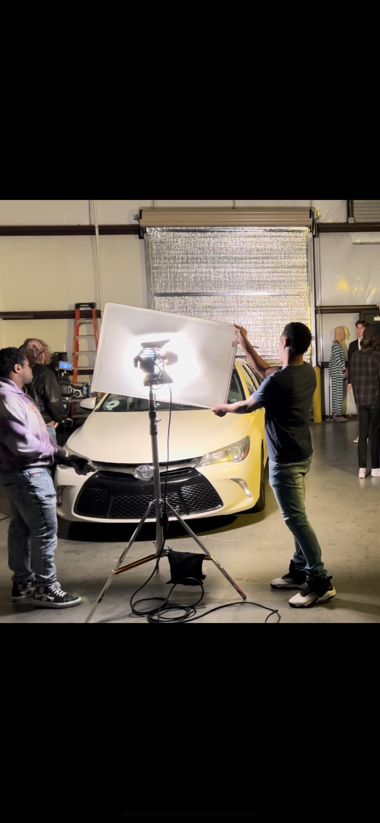 Crew members position a large light and reflector in front of a white car.